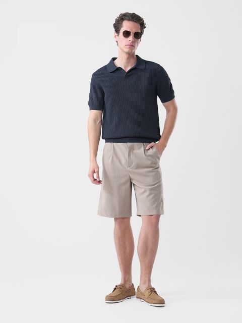 Hombre con gafas de sol, polo azul marino de manga corta, pantalones cortos beige hasta la rodilla y náuticos marrones, de pie frente a un fondo blanco liso.