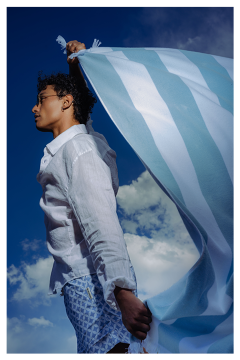 Persona con una camisa blanca, sosteniendo una tela a rayas azules y blancas, frente a un cielo azul con nubes.