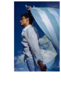 Una persona con una camisa blanca y shorts estampados sostiene una tela a rayas azules y blancas frente a un cielo azul con nubes.