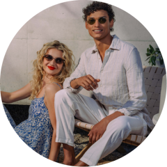 Dos personas con gafas de sol se relajan al aire libre; una está sentada en una silla, la otra está sentada a su lado, ambas sonríen y visten ropa ligera y veraniega.
