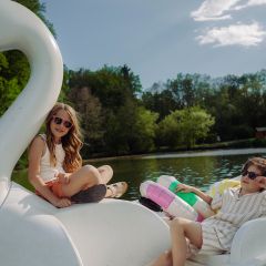 Dos niños con gafas de sol se relajan en un bote de pedales con forma de cisne en un lago, rodeados de árboles bajo un cielo soleado.