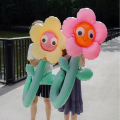 Dos personas sostienen grandes flores inflables con caras sonrientes, que cubren sus propios rostros, mientras están de pie en un puente junto al agua.