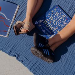 Dos personas están sentadas con las piernas estiradas sobre una manta azul al aire libre, con sandalias y zapatillas deportivas, junto a dos libros abiertos, uno de ellos con una cubierta azul.