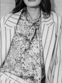 Una mujer con un traje a rayas, con una blusa estampada y pajarita, sonriendo; la foto es en blanco y negro y la muestra de los hombros a la cintura.
