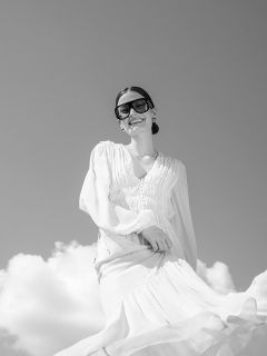 Una mujer con un vaporoso vestido blanco y grandes gafas de sol sonríe bajo un cielo nublado; la foto es en blanco y negro.