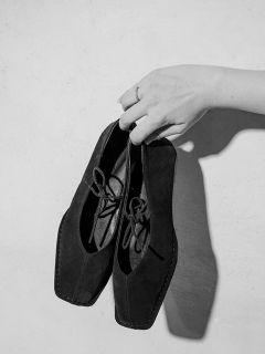 Una mano con un anillo en el dedo sostiene un par de zapatos negros con cordones de punta cuadrada frente a un fondo liso y claro.
