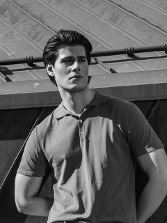 Un hombre con un polo de manga corta está de pie al aire libre, con la mirada hacia un lado, al fondo un tejado metálico inclinado. Foto en blanco y negro.