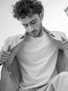 Foto en blanco y negro de un hombre con cabello rizado que mira hacia abajo; lleva una chaqueta ligera sobre una camiseta blanca sencilla y mantiene la chaqueta abierta.