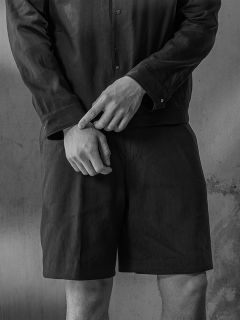 Foto en blanco y negro de una persona en camisa y pantalones cortos, con las manos cruzadas delante de la cintura y de pie frente a una pared texturizada.