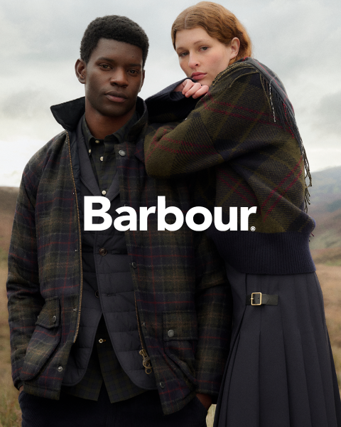 Un hombre y una mujer con chaquetas Barbour a cuadros posan al aire libre, muy juntos, frente a un cielo nublado y un paisaje montañoso. El logo de Barbour está centrado.