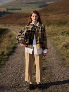 Una persona está de pie en un camino rural en un paisaje de colinas y viste una chaqueta a cuadros, una camisa blanca, unos pantalones beige y zapatos negros.
