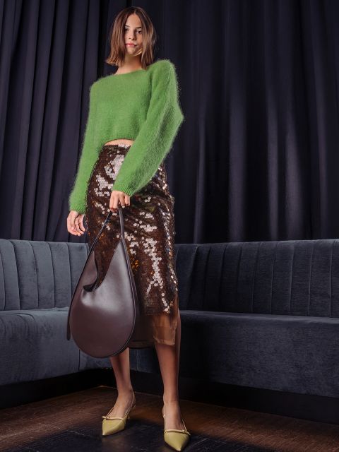 Una mujer posa con un suéter verde esponjoso, una falda midi marrón con lentejuelas y zapatos de tacón amarillos claros, y sostiene un bolso hobo grande de cuero marrón oscuro frente a una cortina oscura.