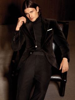 Un hombre con cabello largo y castaño está sentado en un sillón oscuro y lleva puesto un traje de terciopelo negro, un jersey de cuello alto negro y un pañuelo de bolsillo blanco.