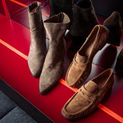 Dos pares de zapatos de ante marrones - botines y mocasines - expuestos en un estante rojo brillante donde se reflejan sus imágenes.
