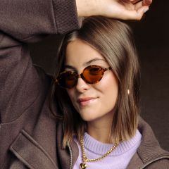 Una mujer con cabello castaño, que lleva gafas de sol con montura de carey, una chaqueta marrón, un top morado y collares dorados, y levanta un brazo sonriendo.
