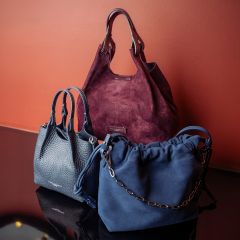 Tres elegantes bolsos de mano - uno en rojo burdeos, uno en azul marino con una cadena y uno en gris oscuro - fueron presentados frente a una pared rojiza sobre una superficie reflectante.