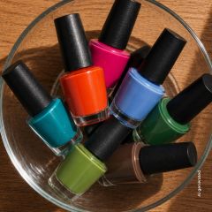 Un cuenco de cristal, lleno de siete frascos de esmalte de uñas en diferentes colores, incluyendo azul, rosa, naranja, verde, verde azulado, verde lima y marrón.
