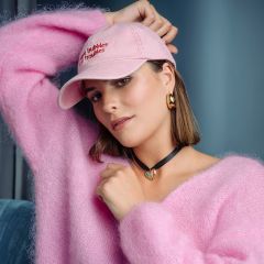 Una mujer con un suéter rosa esponjoso y un gorro rosa posa en una habitación; lleva pendientes dorados y un collar negro con un colgante de corazón turquesa.