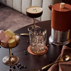 Una mesa con un cóctel, un vaso, un tazón con malvaviscos, una cafetera, una cuchara dorada, una servilleta y granos de café esparcidos.