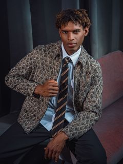 Un hombre con una chaqueta estampada, una camisa y una corbata a rayas está sentado en un sofá rojo frente a una cortina oscura, mirando a la cámara con confianza.