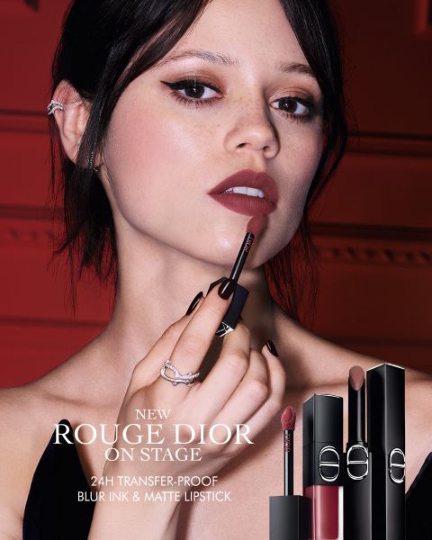 Una mujer se aplica el pintalabios "Rouge Dior" y sostiene el aplicador cerca de sus labios. Se muestran tres tubos de pintalabios. El texto anuncia un pintalabios de 24 horas que no mancha, difumina suavemente los labios y ofrece un acabado mate.