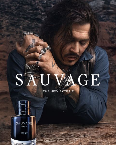 Un hombre con tatuajes y joyas se apoya en una mesa junto a una botella de perfume "Dior Sauvage Extrait" frente a un telón de fondo exterior rugoso.