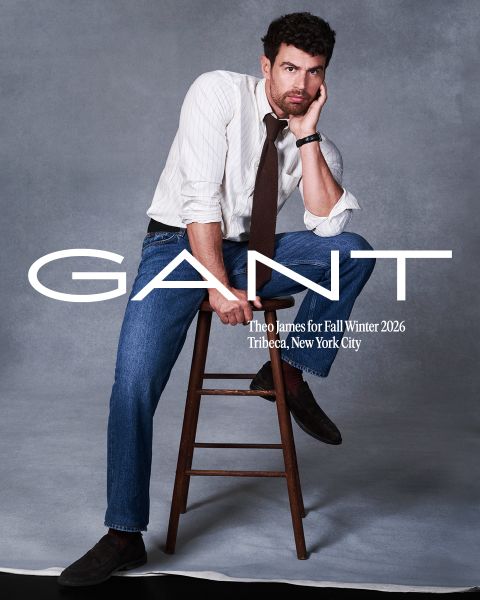 Un hombre con camisa a rayas, corbata y jeans posa en un taburete de madera frente a un fondo gris. Texto: "GANT, Theo James para Otoño/Invierno 2026, Tribeca, Ciudad de Nueva York.".