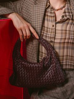 Una persona con un traje a cuadros, sosteniendo un bolso de mano tejido de color marrón oscuro; un fondo rojo se ve parcialmente en el fondo.
