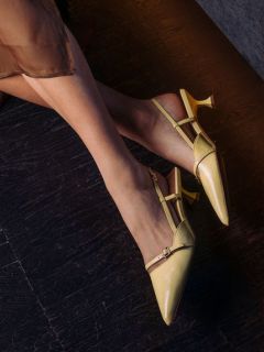 Una mujer con tacones amarillos de punta y tiras está sentada en un suelo de madera oscura; su falda transparente de color marrón es parcialmente visible.