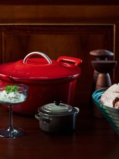 Una olla roja, una olla pequeña gris, un tazón de postre de vidrio, un tazón de pan y molinillos de sal y pimienta sobre una mesa de madera.