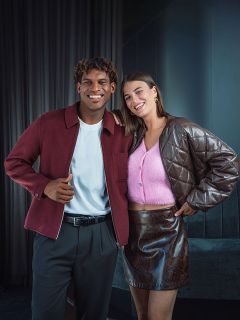 Un hombre sonriente con una chaqueta burdeos está de pie junto a una mujer con un top rosa y una falda marrón; ambos posan en un interior frente a un fondo oscuro.