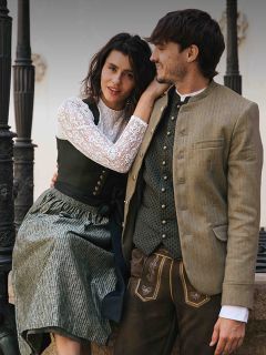 Una mujer y un hombre con trajes bávaros tradicionales están sentados juntos al aire libre, sonríen y se miran.