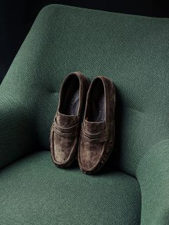 Un par de mocasines de ante marrones yace ordenadamente sobre un sillón tapizado en verde.
