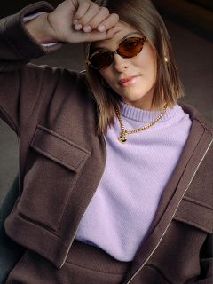 Una mujer con gafas de sol marrones, chaqueta y jersey lavanda apoya el brazo en la frente; lleva joyas de oro y tiene una expresión de confianza.