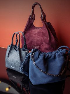 Tres elegantes bolsos de mano - uno de ante burdeos, uno de cuero azul marino y uno de ante azul marino con correa de cadena - están expuestos sobre un fondo marrón rojizo.