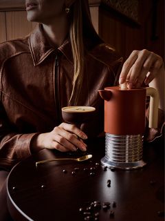 Una mujer con una chaqueta marrón está sentada en una mesa con un cóctel de café, una prensa francesa y granos de café esparcidos.
