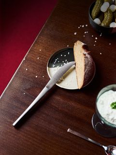 Un cuchillo en un plato con una rebanada de pan, un poco de sal y pepinillos encurtidos, así como una bebida cremosa al lado sobre una mesa de madera.