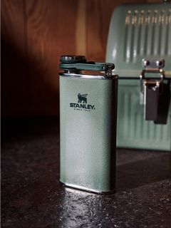 Sobre una encimera oscura hay un termo Stanley verde, y en el fondo, frente a una pared de madera, se ve una fiambrera verde a juego.