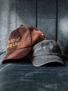 Dos gorras en un sillón de tela azul; una marrón con la inscripción "The Art of Being Late", la otra en negro descolorido con letras más pequeñas y difíciles de leer.