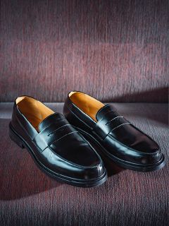 Un par de mocasines de cuero negros brillantes, colocados uno al lado del otro sobre una base de tela texturizada de color burdeos.