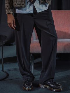 Una persona que lleva pantalones negros anchos, una chaqueta estampada y mocasines negros, está de pie junto a una silla rosa y una mesa redonda pequeña.