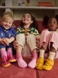 Tres niños pequeños están sentados en un sofá rojo, sonríen y se ríen; cada uno lleva ropa de colores y zapatos diferentes y coloridos.