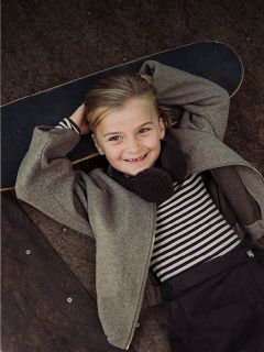 Un niño sonriente con una camisa a rayas y una chaqueta está acostado en una patineta, con las manos entrelazadas detrás de la cabeza, y mira a la cámara.