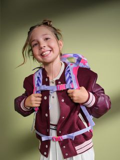 Una chica sonriente con una mochila y una chaqueta color burdeos está de pie frente a un fondo verde, luciendo alegre y lista para la escuela.