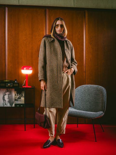 Una persona con un abrigo marrón y una bufanda está de pie en una habitación sobre una alfombra roja, con decoraciones vintage, una silla y una lámpara roja visibles al fondo.