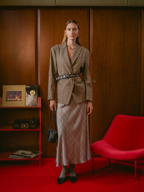 Una mujer en blazer y falda a cuadros está de pie en una habitación retro con paredes de madera, alfombra roja, silla roja y objetos en un estante rojo.