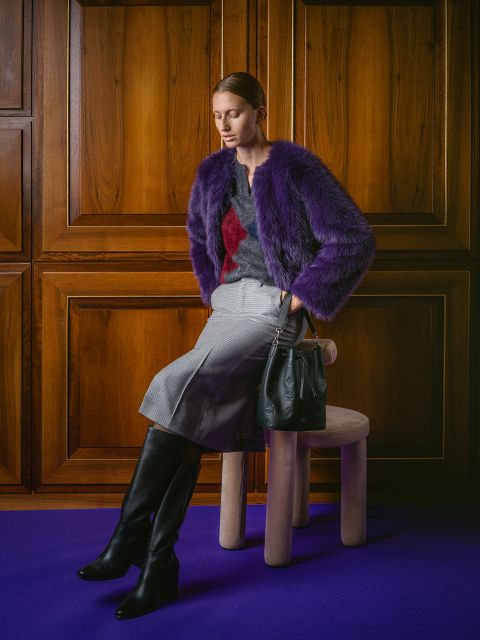 Una mujer con una chaqueta de piel violeta, una falda gris y botas negras está sentada con un bolso negro en un taburete frente a paneles de madera sobre una alfombra violeta.