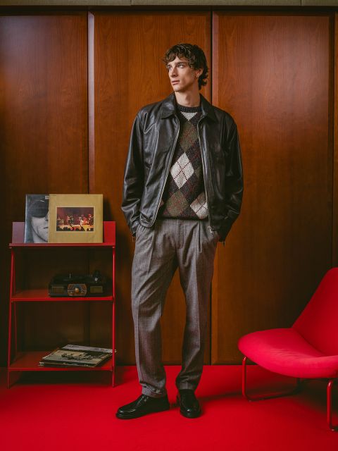 Un hombre con chaqueta de cuero y jersey de cuadros está de pie sobre una alfombra roja junto a un sillón rojo, una radio retro y álbumes frente a paredes con paneles de madera.