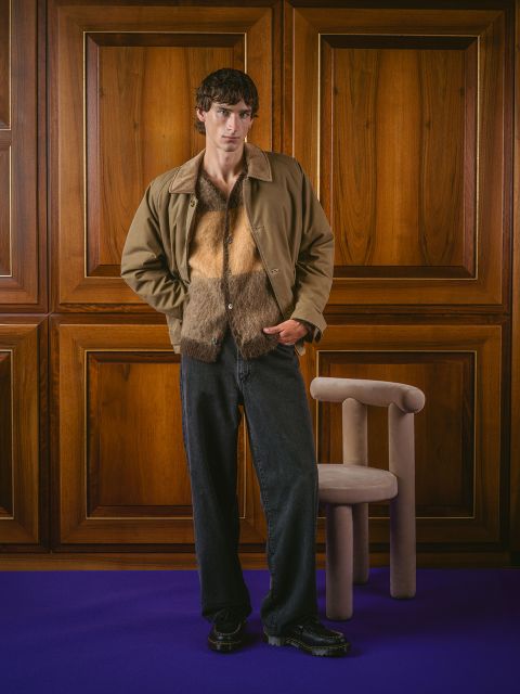 Un hombre con una chaqueta marrón, una camisa peluda y jeans negros está de pie en una habitación junto a un sillón beige, con paredes con paneles de madera y una alfombra morada.