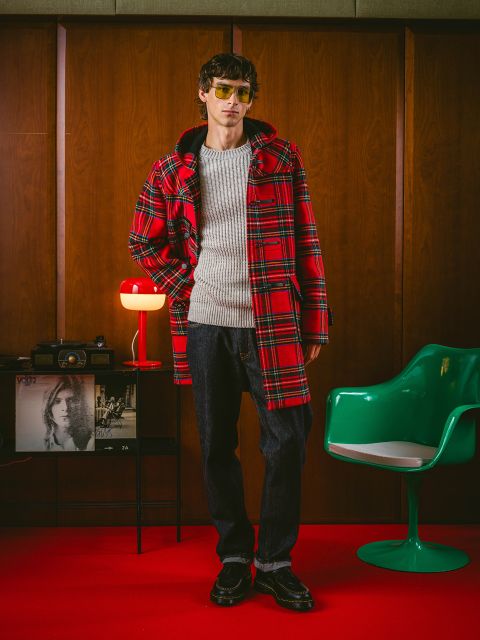 Una persona con un abrigo de cuadros rojo, un suéter gris, jeans oscuros y gafas de sol está de pie en una habitación de estilo retro con paneles de madera, una silla verde y una lámpara roja.